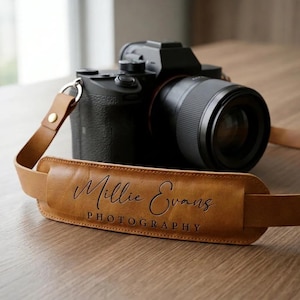May include: A tan leather camera strap featuring a padded shoulder section with the embossed text Millie Evans PHOTOGRAPHY in a combination of script and block lettering, attached to a black digital camera with a lens via metal rings.