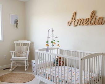 May include: A nursery room featuring a white wooden crib with a patterned sheet, a white rocking chair with a beige cushion, and a large wooden script sign on the wall that reads Amelia. A mobile hangs above the crib.