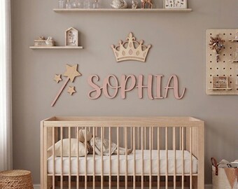Könnte beinhalten: Ein helles Holzbettchen mit weißer Matratze und einer Puppe, ergänzt durch Wanddekoration mit einer Holzkrone, einem Sternenstab und dem Namen SOPHIA in rosa Buchstaben. Zwei Wandregale zeigen kleine Glasflaschen, einen Schwan und ein Reh.