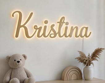 Könnte beinhalten: Ein Wandleuchtschild mit dem Namen Kristina in Schreibschrift und einer warmen, leuchtenden Hintergrundbeleuchtung. Die Buchstaben sind in einem hellen Beigeton gehalten und erzeugen einen sanften Lichtschein an der Wand.