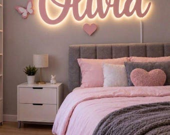 Könnte beinhalten: Ein beleuchtetes Wand-Schild mit dem Namen Olivia in rosa Schreibschrift, verziert mit einer rosa Krone, zwei Schmetterlingen und einem Herz. Das Schild befindet sich über einem Bett mit rosa und grauer Bettwäsche, einem herzförmigen Kissen und einem Nachttisch.