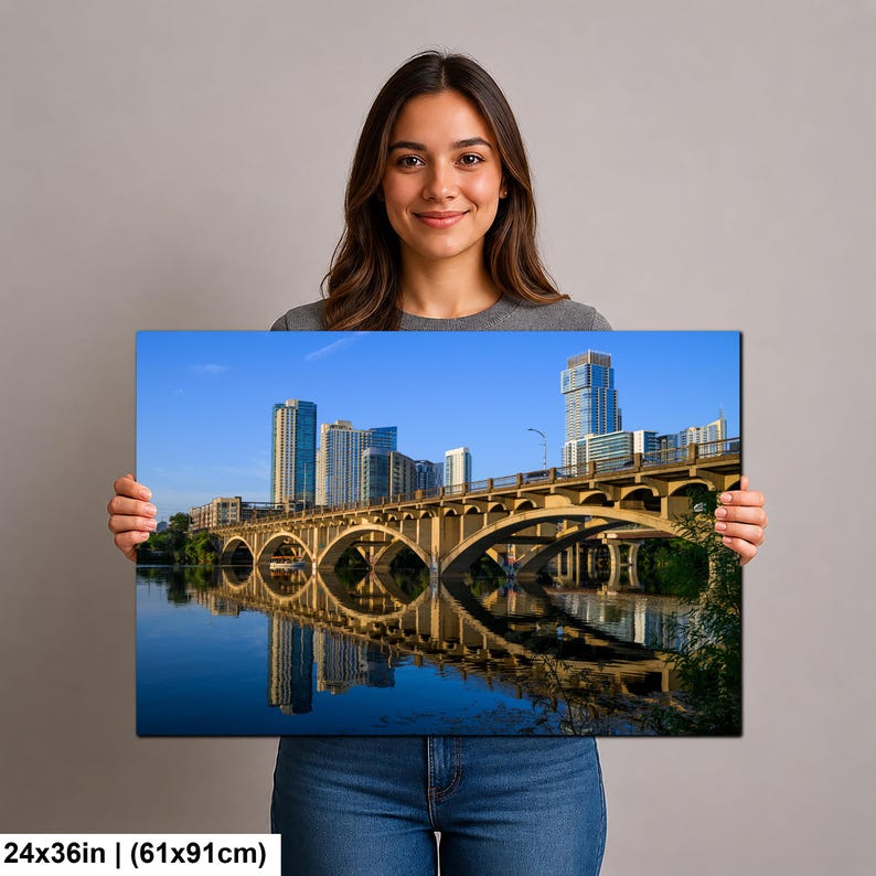 May include: A rectangular wall art print measuring 24 by 36 inches, featuring a vibrant cityscape of Austin, Texas. The image displays the Congress Avenue Bridge with its iconic arches reflecting in the water, set against a clear blue sky.