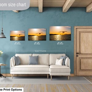 May include: A living room size chart displaying three rectangular wall art prints of a seagull over water at sunset. Sizes include 16x24 inches, 20x30 inches, and 24x36 inches, positioned above a white sectional couch with gray pillows and a side table.