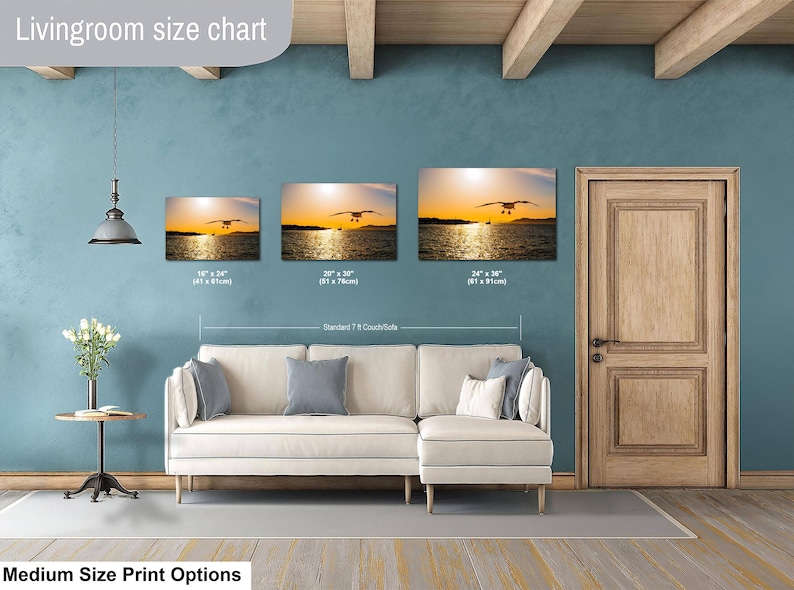 May include: A living room size chart displaying three rectangular wall art prints of a seagull over water at sunset. Sizes include 16x24 inches, 20x30 inches, and 24x36 inches, positioned above a white sectional couch with gray pillows and a side table.