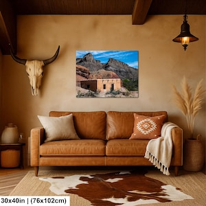 May include: A rectangular wall art print measuring 30 by 40 inches, featuring a landscape photograph of a tan adobe building situated at the base of rugged, dark rocky mountains under a bright blue sky with wispy white clouds.