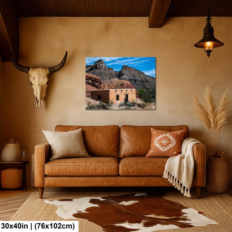May include: A rectangular wall art print measuring 30 by 40 inches, featuring a landscape photograph of a tan adobe building situated at the base of rugged, dark rocky mountains under a bright blue sky with wispy white clouds.