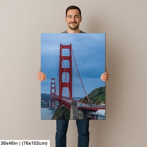May include: A rectangular wall art print featuring the Golden Gate Bridge in San Francisco. The iconic bright red suspension bridge spans across dark blue water under a cloudy sky, with green hills visible in the background. Measures 30x40 inches.