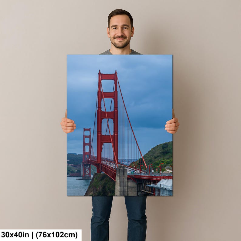 May include: A rectangular wall art print featuring the Golden Gate Bridge in San Francisco. The iconic bright red suspension bridge spans across dark blue water under a cloudy sky, with green hills visible in the background. Measures 30x40 inches.
