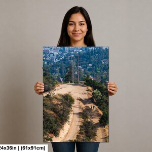 以下が含まれることがあります： 61x91cmの長方形のウォールアートプリント。木々が生い茂る丘陵地帯を曲がりくねって続く土のハイキングコースを高い位置から撮影した風景写真で、澄んだ青空の下、遠くに街並みが見える様子が写し出されています。