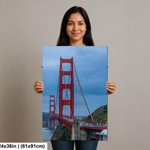 May include: A rectangular wall art print featuring the Golden Gate Bridge in San Francisco, displaying its iconic red-orange towers and suspension cables against a cloudy blue sky, measuring 24 inches by 36 inches in total dimensions.
