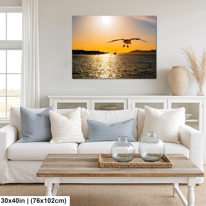 May include: A rectangular wall art print measuring 30x40 inches featuring a silhouette of a seagull in flight over a body of water at sunset. The golden sun hangs low in the sky above distant hills and a small boat, with light reflecting on the water.