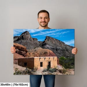 May include: A rectangular wall art print measuring 30 by 40 inches, featuring a landscape photograph of a tan adobe building situated at the base of rugged, dark gray rocky mountains under a bright blue sky with scattered white clouds.