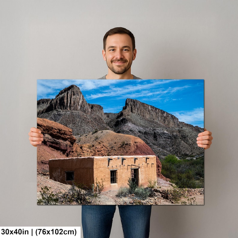 May include: A rectangular wall art print measuring 30 by 40 inches, featuring a landscape photograph of a tan adobe building situated at the base of rugged, dark gray rocky mountains under a bright blue sky with scattered white clouds.