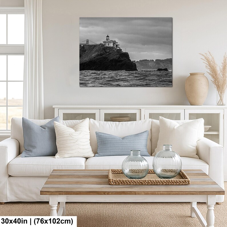 May include: A black and white wall art print depicting a lighthouse perched on a rocky cliff overlooking the ocean under a cloudy sky. The artwork measures 30 inches by 40 inches and is displayed above a white sofa with decorative pillows.