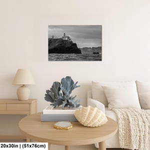 May include: A black and white wall art print depicting a lighthouse on a rocky cliff overlooking the ocean under a cloudy sky. The print measures 20 by 30 inches and is displayed on a wall above a wooden table with a lamp and decor.