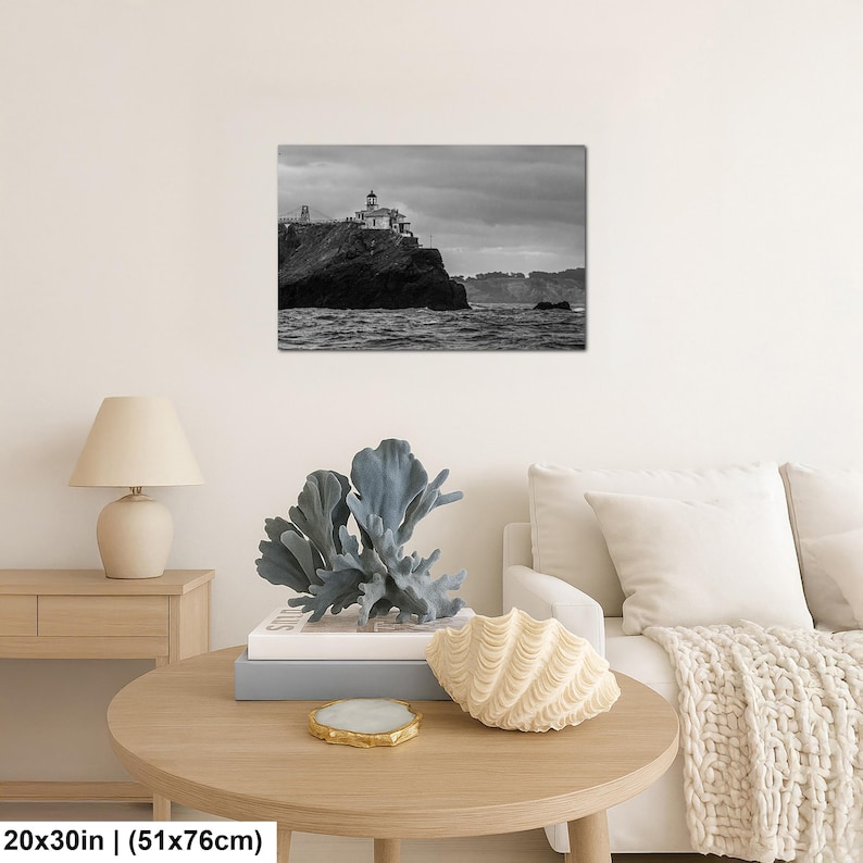 May include: A black and white wall art print depicting a lighthouse on a rocky cliff overlooking the ocean under a cloudy sky. The print measures 20 by 30 inches and is displayed on a wall above a wooden table with a lamp and decor.