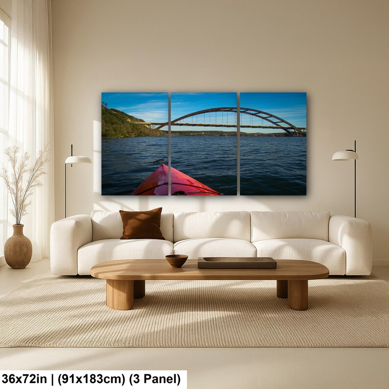 May include: A three-panel wall art set depicting a view from a red kayak on dark blue water, looking toward a large arched bridge under a bright blue sky. The artwork measures 36 by 72 inches and features a landscape scene with a shoreline.