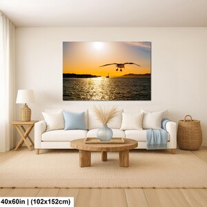 May include: A rectangular wall art print featuring a seagull in flight over a body of water at sunset, displayed above a white sofa with light blue and white pillows, a wooden coffee table, a lamp, and a woven basket, measuring 40x60 inches.