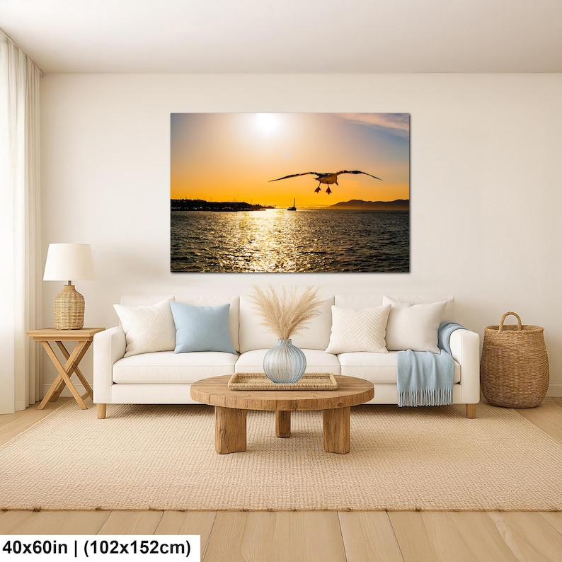 May include: A rectangular wall art print featuring a seagull in flight over a body of water at sunset, displayed above a white sofa with light blue and white pillows, a wooden coffee table, a lamp, and a woven basket, measuring 40x60 inches.