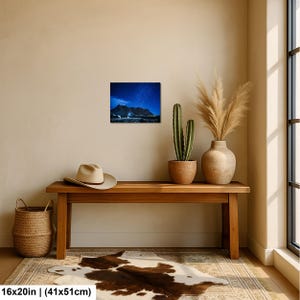 Puede incluir: Una impresi&oacute;n de arte mural rectangular de 41 por 51 cm, que muestra un paisaje nocturno de una cordillera oscura bajo un cielo estrellado azul profundo. La impresi&oacute;n se exhibe en una pared lisa sobre un banco de madera, un sombrero y una cesta.