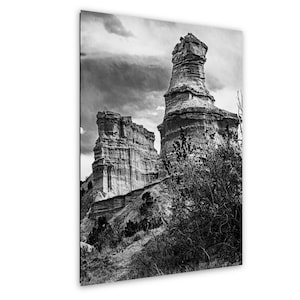 May include: A black and white photograph of tall, weathered rock formations rising from a rugged landscape. The scene features layered stone pillars under a cloudy sky, with dark, dense shrubbery in the foreground adding depth to the view.