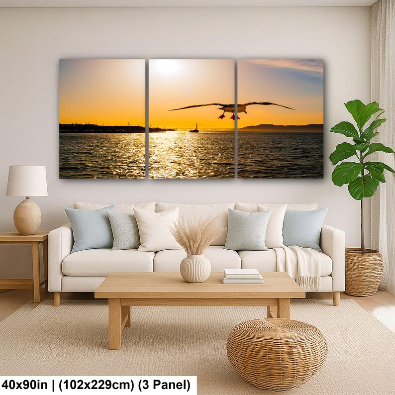 May include: A three-panel wall art set depicting a seagull in flight over a body of water at sunset with a golden sky and distant shoreline. The artwork measures 40 by 90 inches and is displayed above a sofa in a living room setting.