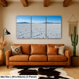 May include: A three-panel wall art set depicting a vast white salt flat landscape under a clear blue sky with distant mountains. The artwork measures 36 by 72 inches and is displayed above a brown leather sofa in a living room setting.