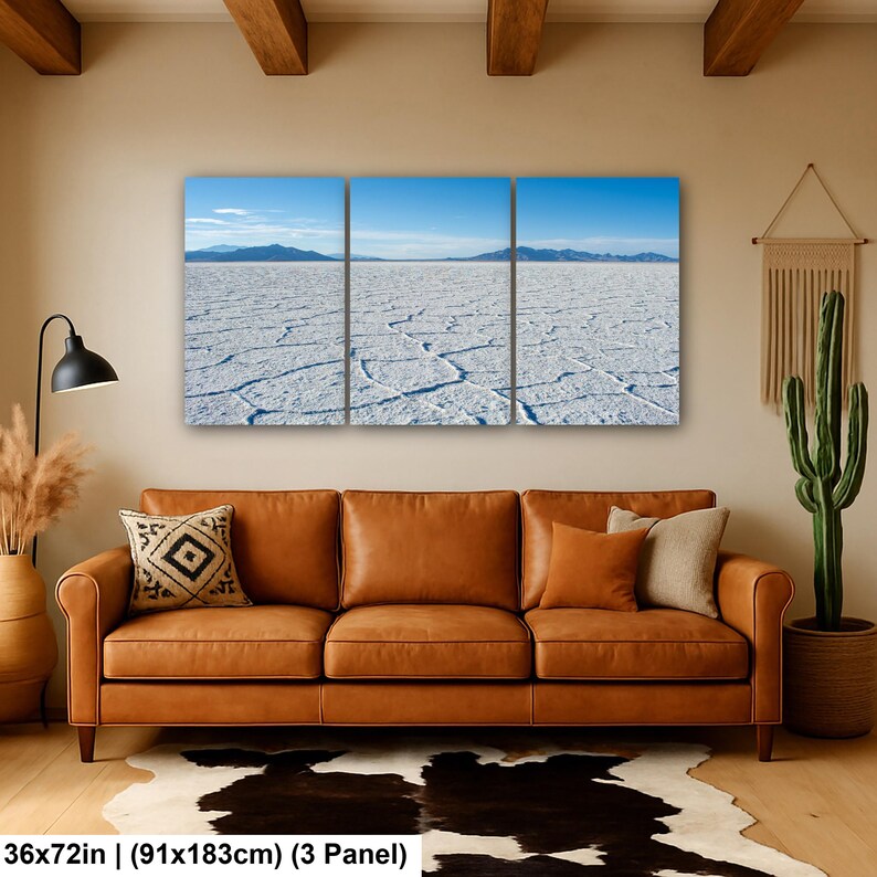 May include: A three-panel wall art set depicting a vast white salt flat landscape under a clear blue sky with distant mountains. The artwork measures 36 by 72 inches and is displayed above a brown leather sofa in a living room setting.