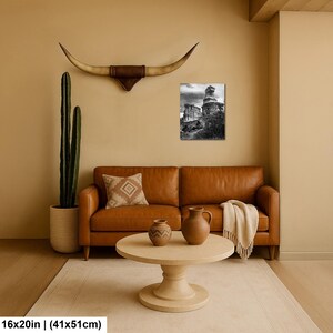 May include: A black and white wall art print depicting a rocky landscape, mounted above a brown leather sofa. The scene includes a tall cactus in a textured pot, a longhorn steer skull wall mount, and a round wooden coffee table with two vases.
