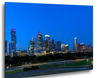 May include: A rectangular wall art print featuring a twilight cityscape of Austin, Texas. The image displays illuminated skyscrapers against a deep blue sky, with a green park and paved pathways visible in the foreground under soft lighting.