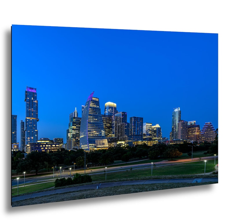 A rectangular wall art print featuring a twilight cityscape of Austin, Texas. The image displays illuminated skyscrapers against a deep blue sky, with a green park and paved pathways visible in the foreground under soft street lamp lighting.