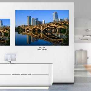 May include: Two rectangular wall art prints featuring a city skyline and a bridge over water. The smaller print is 30 x 40 inches and the larger print is 40 x 60 inches. The prints are displayed above a white reception desk in an office space.