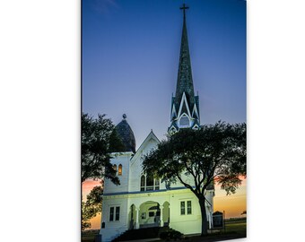 May include: A white wooden church building with a tall steeple topped by a cross, featuring arched windows and a covered entrance. The structure is illuminated against a twilight sky with orange and blue hues, framed by dark green trees.