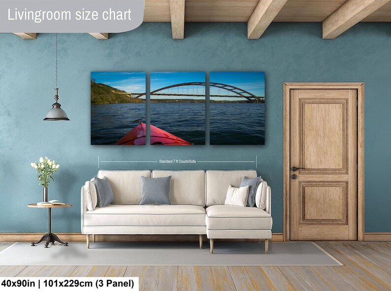 May include: A three-panel canvas wall art set depicting a red kayak on a lake facing a large arched bridge under a blue sky. The artwork is displayed above a cream-colored sectional sofa. The text Livingroom size chart and 40x90in | 101x229cm (3 Panel) is visible.