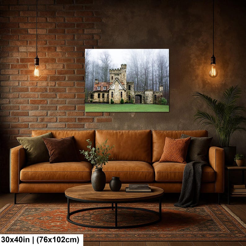 Op de afbeelding: Een rechthoekige wandkunstprint met een stenen kasteel met een zadeldak tegen een achtergrond van een mistig bos. De print meet 76 cm bij 102 cm en hangt aan een bakstenen muur boven een bruine leren bank.