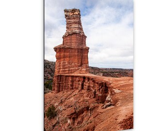 May include: A tall, vertical red rock formation stands prominently against a cloudy sky. The geological structure features layered, textured surfaces with deep orange and brown hues, rising from a rugged, arid landscape of eroded canyon terrain.