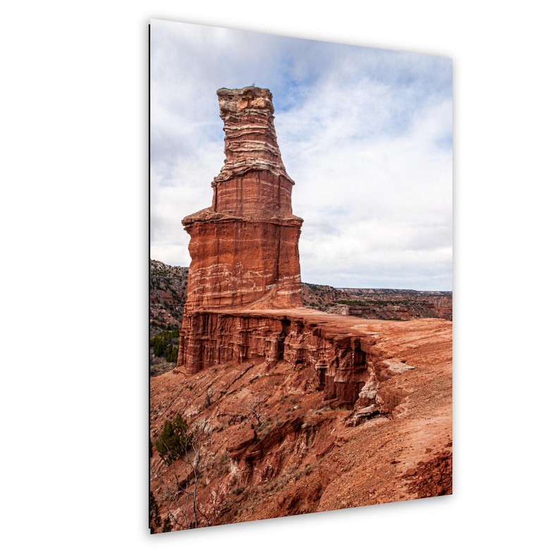 May include: A tall, vertical red rock formation stands prominently against a cloudy sky. The geological structure features layered, textured surfaces with deep orange and brown hues, rising from a rugged, arid landscape of eroded canyon terrain.