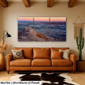 May include: A three-panel wall art set depicting a desert landscape at sunset with layered rock formations in shades of orange, blue, and purple. The artwork measures 36 by 72 inches and features a horizon line transitioning from pink to deep blue.