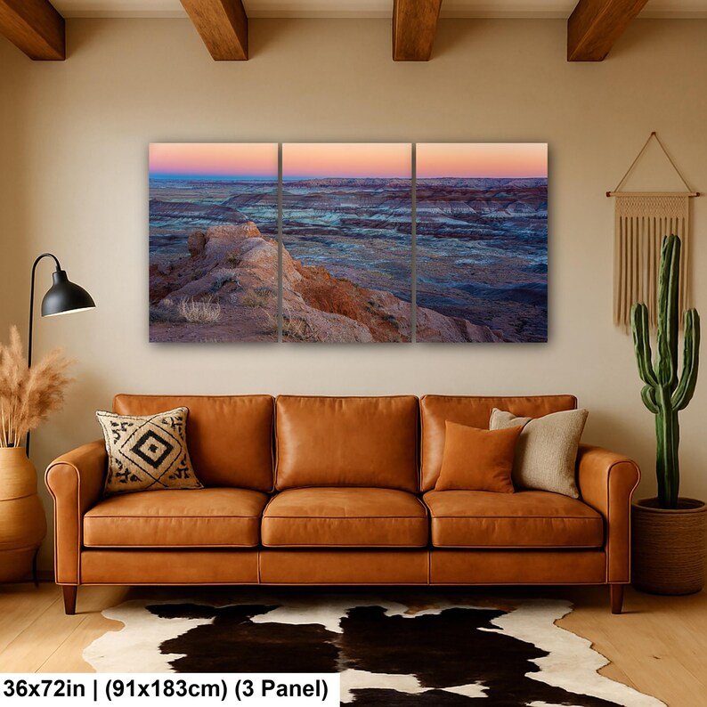 May include: A three-panel wall art set depicting a desert landscape at sunset with layered rock formations in shades of orange, blue, and purple. The artwork measures 36 by 72 inches and features a horizon line transitioning from pink to deep blue.