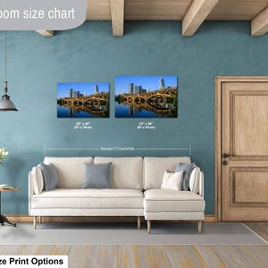 May include: A living room size chart displaying two rectangular wall art prints of a city bridge over water, labeled 20 x 30 inches and 24 x 36 inches, positioned above a cream-colored sectional sofa with gray pillows and a small side table.