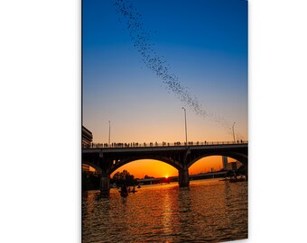 May include: A vertical wall art print depicting a bridge over a river at sunset. A large swarm of bats flies in a diagonal line across the blue and orange sky. Silhouettes of people stand on the bridge, and small boats are visible on the water.