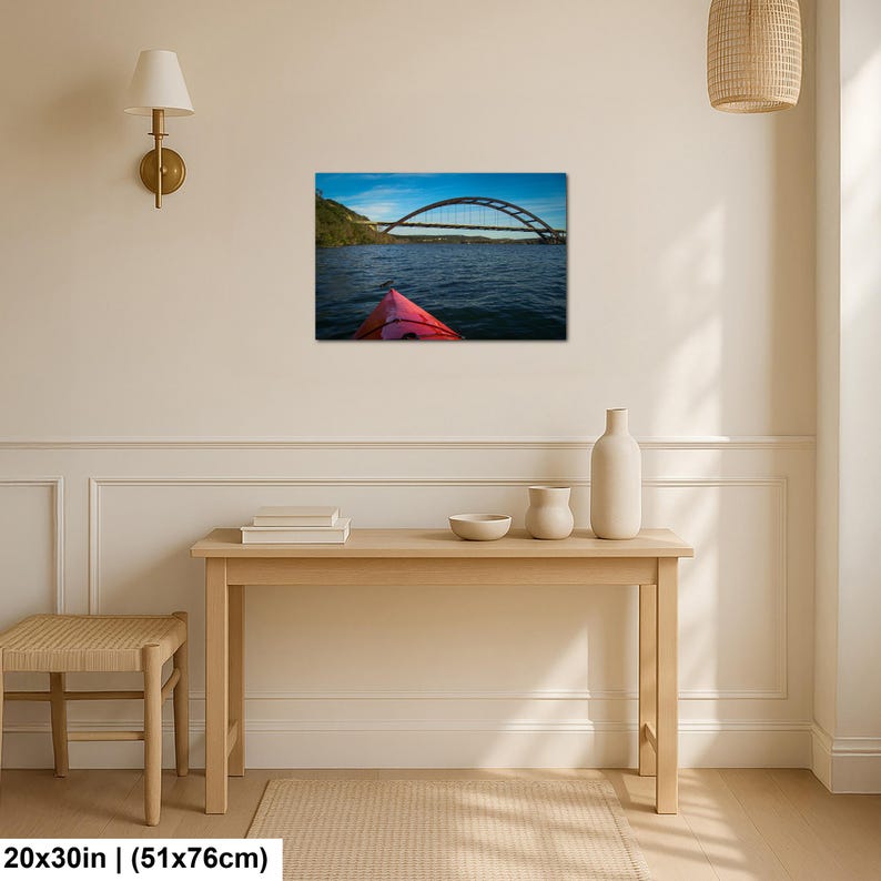 May include: A rectangular wall art print measuring 20 by 30 inches, featuring a landscape photograph of a red kayak bow on dark blue water, looking toward a large arched bridge under a bright blue sky with green hills in the distance.