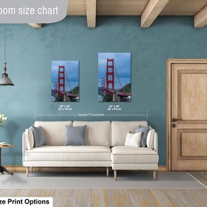 May include: A living room size chart displaying two vertical canvas prints of the Golden Gate Bridge in red, with sizes 20 x 30 inches and 24 x 36 inches, positioned above a cream-colored sectional sofa against a blue wall with a wooden door.