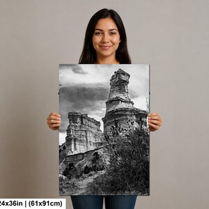 May include: A black and white rectangular wall art print measuring 24 by 36 inches, featuring a landscape of tall, weathered rock formations under a cloudy sky, with sparse desert vegetation in the foreground and a rocky terrain base.