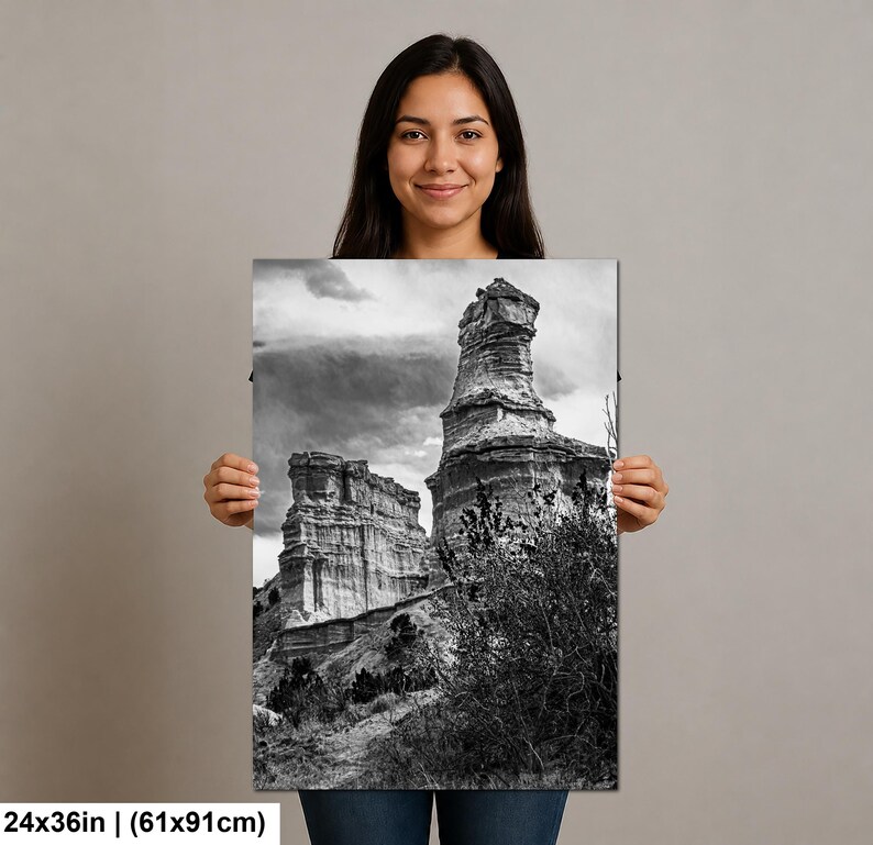May include: A black and white rectangular wall art print measuring 24 by 36 inches, featuring a landscape of tall, weathered rock formations under a cloudy sky, with sparse desert vegetation in the foreground and a rocky terrain base.
