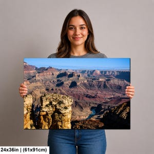May include: A rectangular wall art print featuring a panoramic landscape of the Grand Canyon with layered rock formations in shades of brown, orange, and red, a winding blue river below, and a clear blue sky. The print measures 24x36 inches.