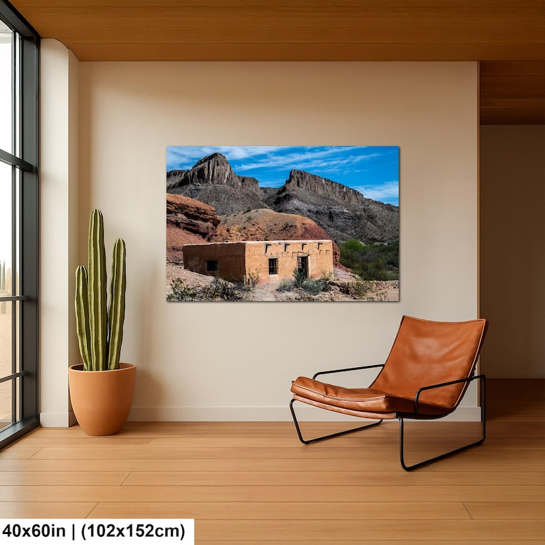 May include: A rectangular wall art print measuring 40 by 60 inches, featuring a landscape photograph of a tan adobe building set against rugged, dark rocky mountains under a bright blue sky, displayed in a room with a cactus and leather chair.