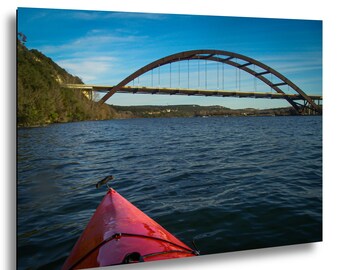 May include: A bright red kayak bow points toward a large steel arch bridge spanning a wide river. The water is dark blue with gentle ripples, and the sky above is a clear, vibrant blue. A steep, tree-covered hillside lines the riverbank.