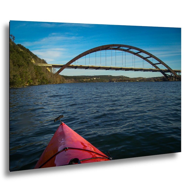 May include: A bright red kayak bow points toward a large steel arch bridge spanning a wide river. The water is dark blue with gentle ripples, and the sky above is a clear, vibrant blue. A steep, tree-covered hillside lines the riverbank.