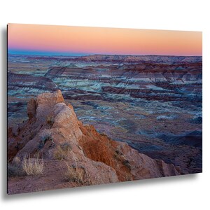 May include: A landscape photograph of a desert badlands terrain featuring layered sedimentary rock formations in shades of red, orange, and blue. The horizon is illuminated by a soft sunset sky with gradients of pink, orange, and deep blue.